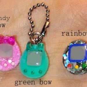 May include: Three colourful, glittery, handheld game keychains. The keychains are in the shape of a classic virtual pet device. One is pink with a bow, one is green with a bow, and the third is rainbow-coloured. The text "candy bow", "green bow", and "rainbow" are visible.