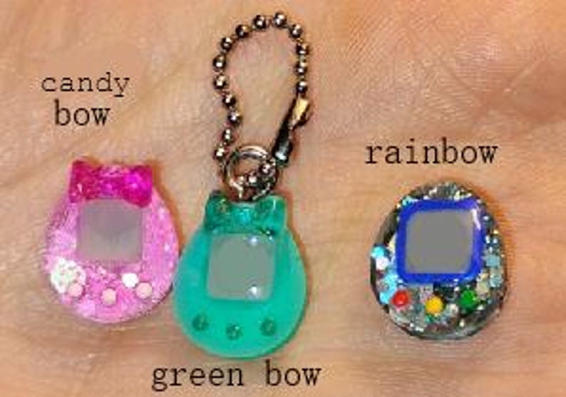 May include: Three colourful, glittery, handheld game keychains. The keychains are in the shape of a classic virtual pet device. One is pink with a bow, one is green with a bow, and the third is rainbow-coloured. The text "candy bow", "green bow", and "rainbow" are visible.