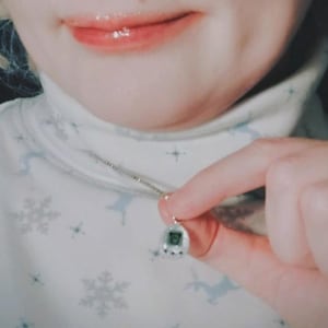 May include: Silver necklace featuring a small, retro pendant with a digital display and buttons. The person is wearing a white turtleneck with a snowflake and deer pattern.