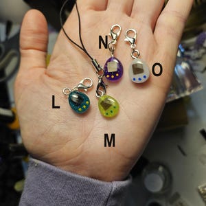 May include: Four colourful, miniature charm pendants in the shape of a retro handheld game. The pendants are teal, green, purple, and white, with yellow and blue accents. Each charm has a silver clasp and a black cord.