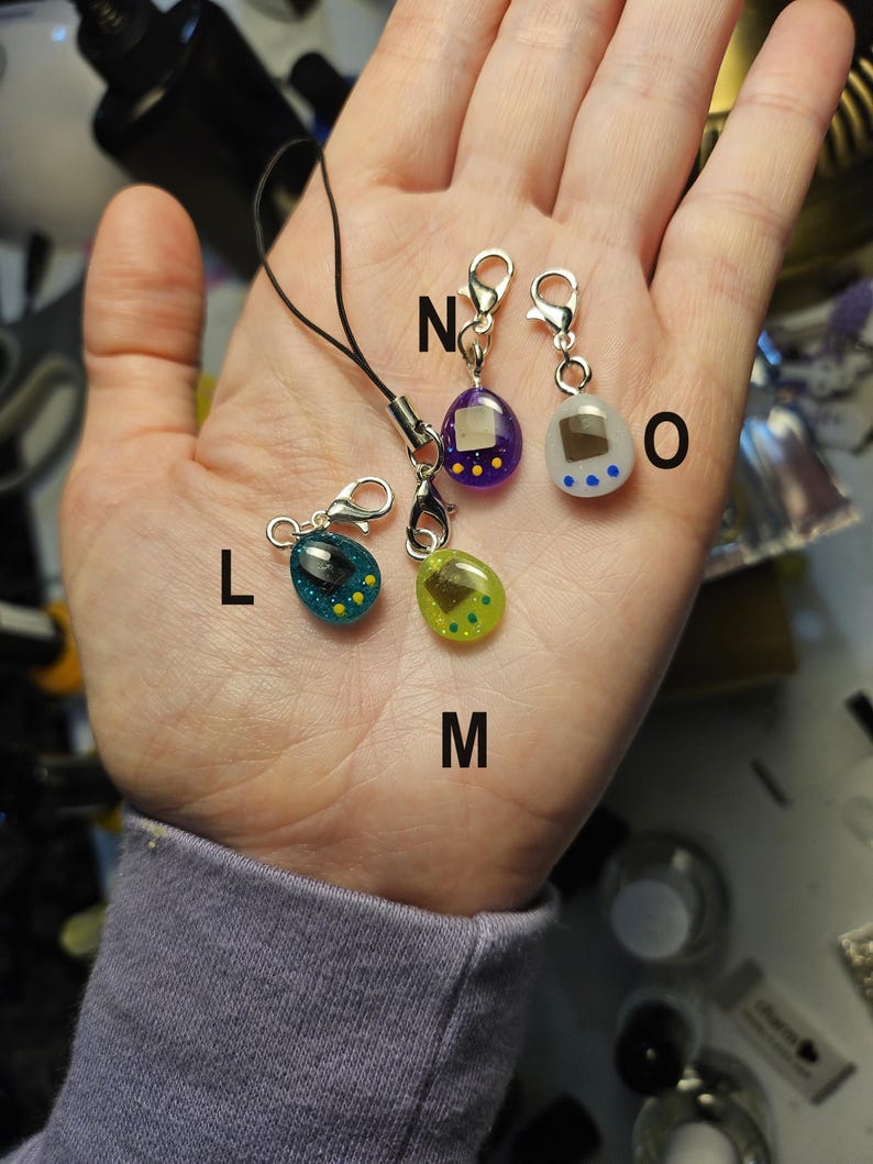 May include: Four colourful, miniature charm pendants in the shape of a retro handheld game. The pendants are teal, green, purple, and white, with yellow and blue accents. Each charm has a silver clasp and a black cord.