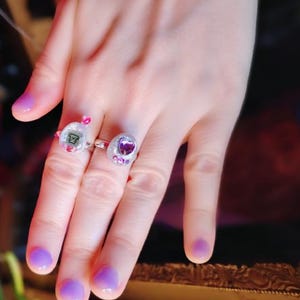 May include: Two rings with clear settings are displayed on a hand with purple nail polish. One ring features a small bear graphic, the other a purple heart. Both rings have pink and purple accents, creating a playful and whimsical aesthetic.