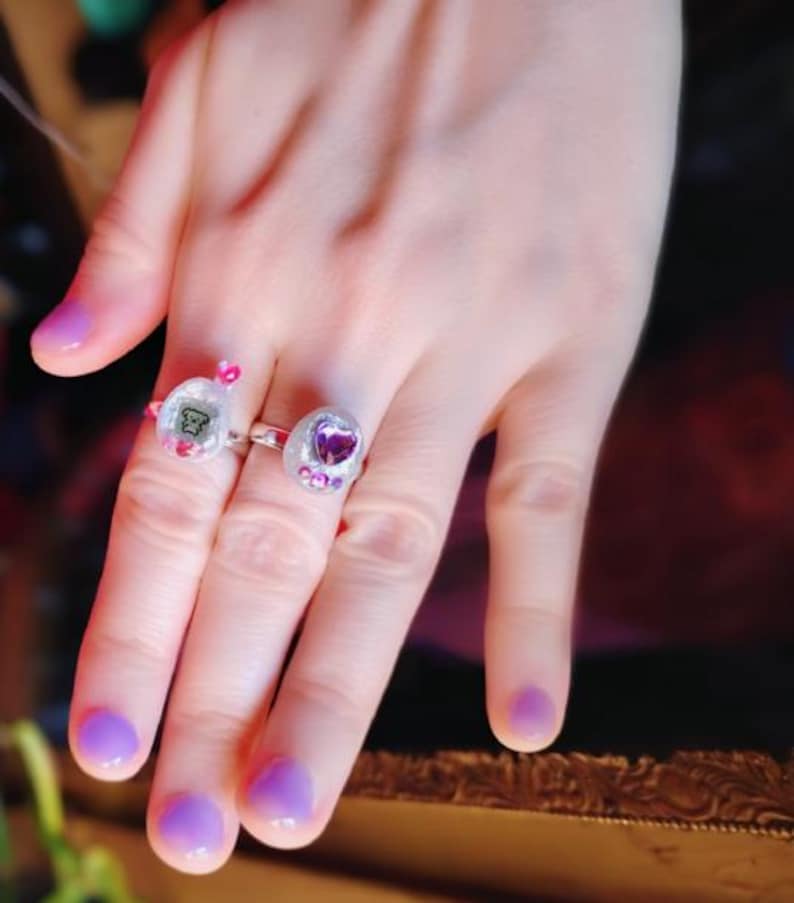 May include: Two rings with clear settings are displayed on a hand with purple nail polish. One ring features a small bear graphic, the other a purple heart. Both rings have pink and purple accents, creating a playful and whimsical aesthetic.