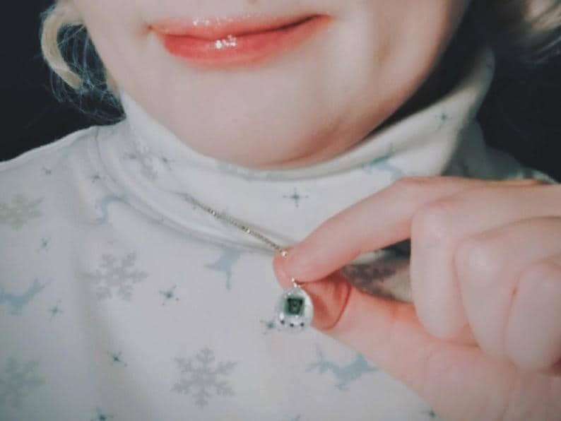 May include: Silver necklace with a small, retro-style pendant. The pendant has a digital display and a button. The person is wearing a white turtleneck with a snowflake and deer pattern.