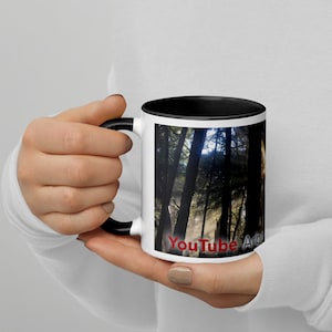 Puede incluir: Taza de cerámica blanca con borde y asa negros. La taza presenta una foto de un bosque con la luz del sol brillando a través de los árboles. El texto "YouTube Arbo" está impreso en la parte inferior de la taza.