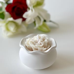 Peut inclure: Une bougie blanche en forme de rose dans un pot en céramique blanc à bords festonnés. Une rose rouge et des lys blancs sont flous en arrière-plan. La bougie a une seule mèche.