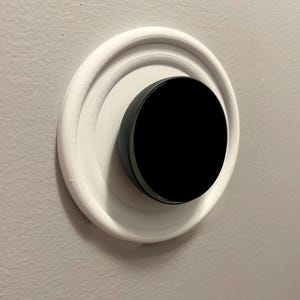Puede incluir: Un termostato inteligente negro y circular montado sobre una base blanca y circular. La base está fijada a una pared gris claro. El termostato tiene un acabado liso y brillante.