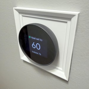 Puede incluir: Un termostato inteligente con una pantalla circular negra, configurado a 60 grados, montado en un marco rectangular blanco. La pantalla muestra "Heat set to 60 Indoor 76". El marco está montado en una pared gris.