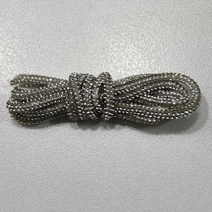 May include: A silver-toned shoelace covered in small, reflective, bead-like embellishments. The cord is coiled into a horizontal, elongated shape, showcasing a uniform, textured surface throughout its entire length with a metallic, shimmering finish.