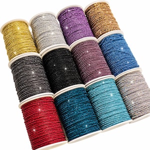 Puede incluir: Una colección de doce carretes cilíndricos envueltos en adornos de diamantes de imitación brillantes, con una variedad de colores que incluyen oro, plata, morado, bronce, negro, rosa oscuro, azul, rojo, verde azulado y blanco iridiscente.