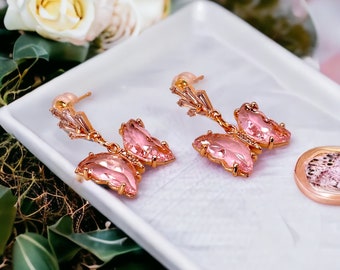 May include: A pair of rose gold butterfly earrings with pink crystal wings. The earrings are displayed on a white tray with a blurred floral background. The earrings have a delicate, feminine design.