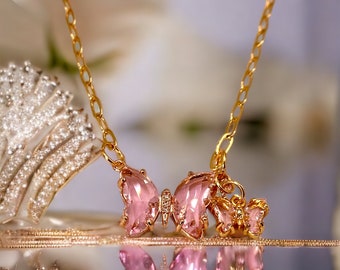 May include: A gold chain necklace with a pink crystal butterfly pendant. The butterfly has two smaller butterflies attached to it.