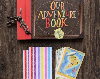 May include: A rectangular scrapbook titled Our Adventure Book with a dark brown cover, red spine, and brown rope binding. Included are two sheets of colorful vertical striped stickers and a stack of postcards featuring a map and airship illustration.