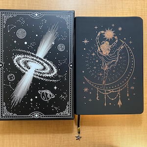 May include: Two black hardcover journals with gold foil accents. The journal on the left has a galaxy design with white stars and a white spiral galaxy. The journal on the right has a gold foil design of a woman dancing on a crescent moon with stars and other celestial elements.