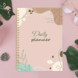 May include: A pink and brown daily planner with a gold spiral binding. The cover features abstract shapes and a leafy design. The text "Daily planner" is written in brown cursive.