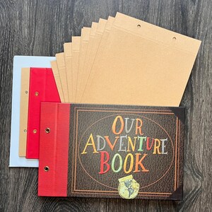 May include: A scrapbook with a dark brown textured cover featuring the text Our Adventure Book in colorful letters and a small globe icon. The book includes a red spine with metal grommets and multiple loose internal pages in tan and red colors.
