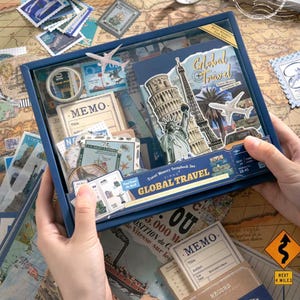 May include: A blue boxed travel memory scrapbook set featuring a notebook, stickers, and memo cards. The box cover displays the text Global Travel and illustrations of the Statue of Liberty, the Eiffel Tower, and an airplane on a map background.