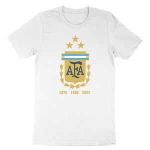 Op de afbeelding: Wit T-shirt met een gouden schild met een blauw "AFA"-logo, drie gouden sterren en een blauw-witte streep. Het schild is omgeven door gouden laurierbladeren. De tekst "1978 1986 2022" is onder het schild gedrukt.