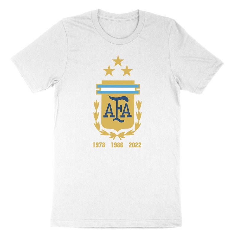 Op de afbeelding: Wit T-shirt met een gouden schild met een blauw "AFA"-logo, drie gouden sterren en een blauw-witte streep. Het schild is omgeven door gouden laurierbladeren. De tekst "1978 1986 2022" is onder het schild gedrukt.