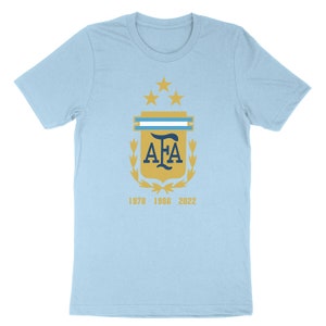 Op de afbeelding: Lichtblauw T-shirt met een gouden wapenschild met de letters "AFA" in donkerblauw, omringd door een lauwerkrans en vijf gouden sterren. De jaren "1978", "1986" en "2022" zijn onder het wapenschild gedrukt.