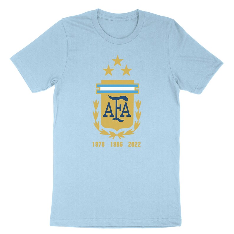 Op de afbeelding: Lichtblauw T-shirt met een gouden wapenschild met de letters "AFA" in donkerblauw, omringd door een lauwerkrans en vijf gouden sterren. De jaren "1978", "1986" en "2022" zijn onder het wapenschild gedrukt.