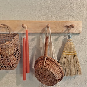 May include: A wooden wall rack with three hooks holds a wicker basket filled with items, a woven basket with a handle, and a straw broom with a yellow stripe.