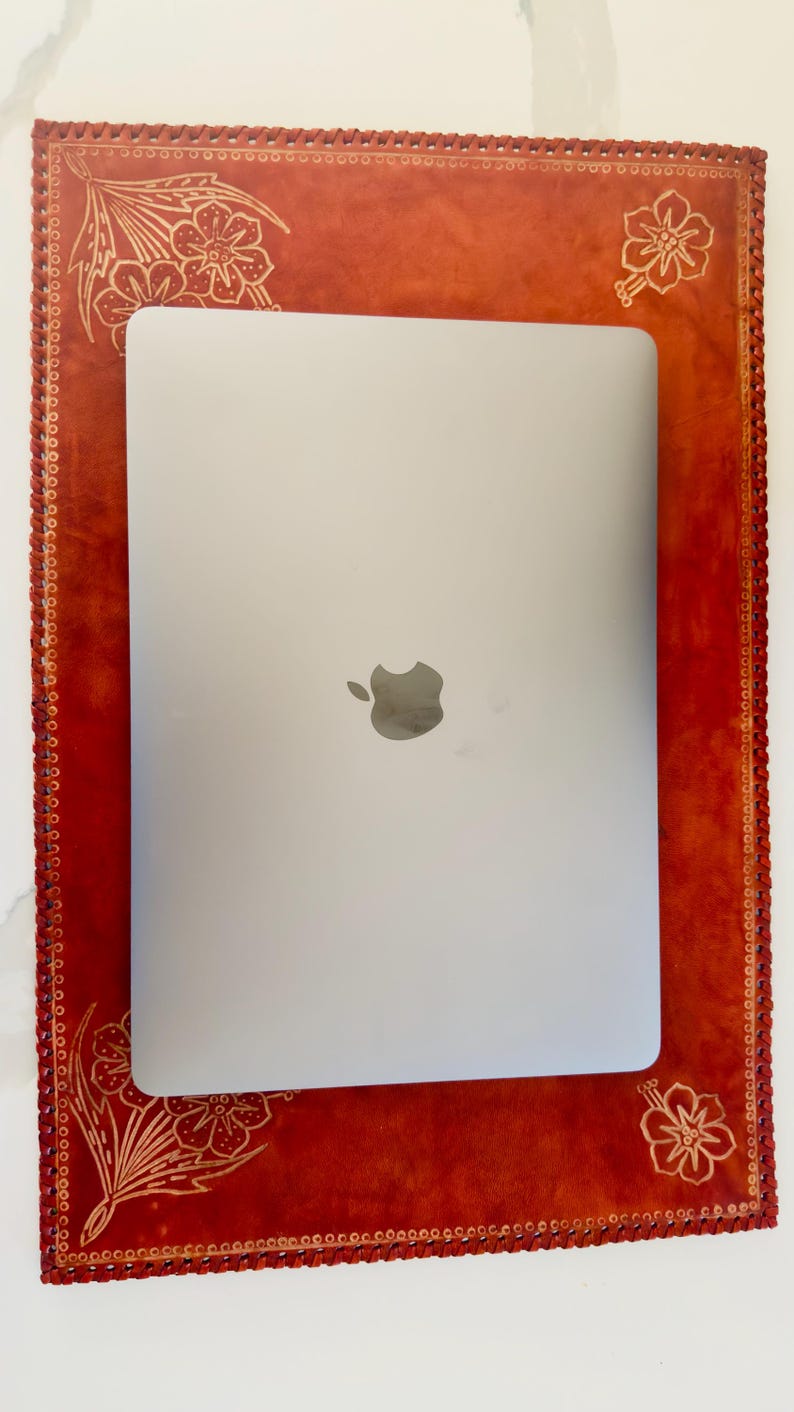 May include: A red leather laptop case with a decorative floral design. The case has a stitched border and a cutout for a laptop.