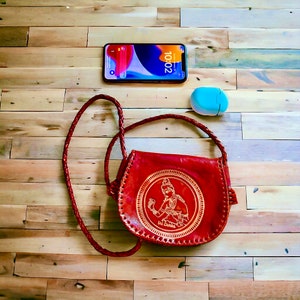 May include: A red leather crossbody bag with a gold embossed design featuring a woman in traditional clothing. The bag has a long strap and a flap closure. The bag is sitting on a wooden surface with a blue phone and a white case.