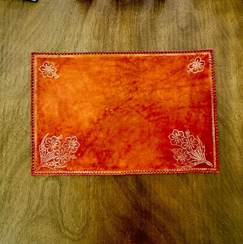 May include: Orange leather placemat with a decorative border and white floral designs.
