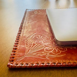 May include: A brown leather laptop desk pad with a floral design and a braided edge. The pad is on a wooden table.