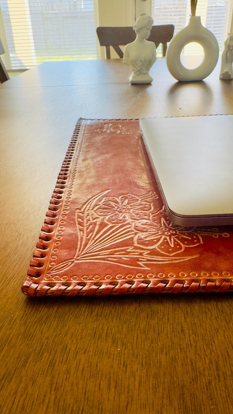May include: A brown leather laptop desk pad with a floral design and a braided edge. The pad is on a wooden table.