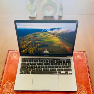 May include: A silver laptop computer with a bright green and brown landscape on the screen. The laptop is sitting on a red leather desk mat with a floral design. The time on the screen is 3:55.