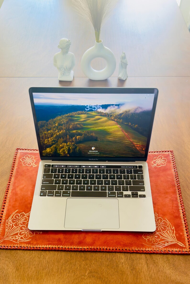 May include: A silver laptop computer with a bright green and brown landscape on the screen. The laptop is sitting on a red leather desk mat with a floral design. The time on the screen is 3:55.