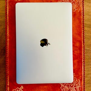 May include: A white laptop computer with the Apple logo on the lid is sitting on a red leather desk pad with white floral designs. The desk pad has a decorative stitched border.