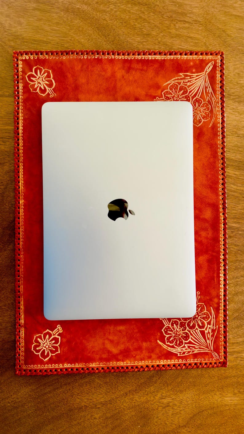May include: A white laptop computer with the Apple logo on the lid is sitting on a red leather desk pad with white floral designs. The desk pad has a decorative stitched border.