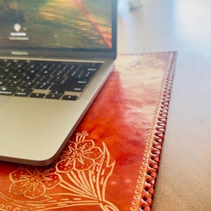 May include: A red leather laptop desk pad with a floral design. The pad has a stitched edge and is made of genuine leather.