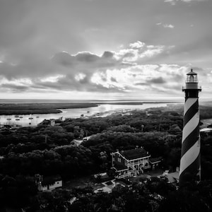 May include: A black and white aerial view of a lighthouse with a striped design. The lighthouse is located on a hill overlooking a body of water and a small town.