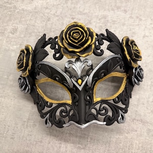 Può includere: Una maschera di carnevale nera e oro con accenti di rose. La maschera presenta un design floreale con dettagli dorati e argentati. Le aperture degli occhi sono bordate d'oro. La maschera è progettata per un evento formale.