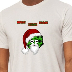 Puede incluir: Camiseta blanquecina con un gráfico de Papá Noel verde con un gorro rojo y barba blanca. Las palabras "HIGH! HIGH! HIGH!" están impresas encima del gráfico en rojo y verde. La camiseta la lleva una persona.