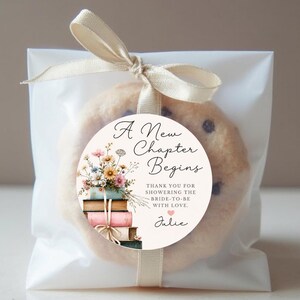 May include: A chocolate chip cookie in a clear cellophane bag, tied with a cream ribbon. A round sticker on the cookie reads "A New Chapter Begins" with floral and book illustrations. Text includes "Thank you for showering the bride-to-be with love."