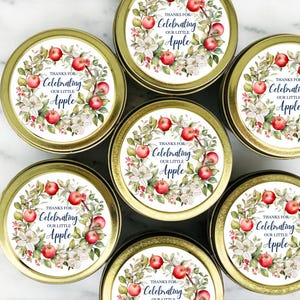 May include: Seven round, gold-colored tins with lids. Each lid features a circular label with a wreath of red apples, white flowers, and green leaves. The text on the label reads "Thanks for Celebrating our little Apple."