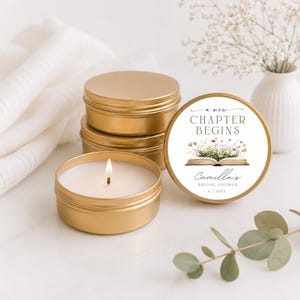 May include: A gold metal candle tin with a white wax candle and a lit wick. A matching gold lid features a label with the text A NEW CHAPTER BEGINS, an illustration of an open book with wildflowers, and the text Camilla's BRIDAL SHOWER 6.2.20XX.