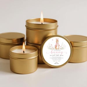 May include: A collection of gold-colored metal candle tins, some with lit wicks. One tin features a circular label with a watercolor illustration of a rabbit, floral accents, and the text A LITTLE BUNNY IS ON THE WAY, followed by a poem.