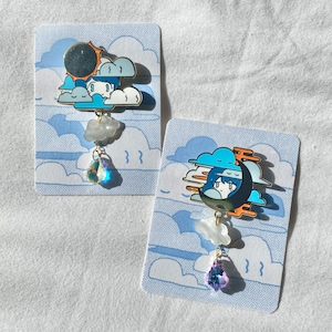 May include: Two enamel pins featuring a blue and white cartoon character with a crescent moon and clouds. The pins are attached to a white card with a blue cloud pattern. The pins have a crystal charm hanging from them.
