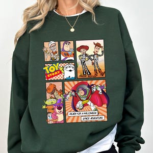 May include: Dark green sweatshirt featuring a colorful comic strip design with characters from the Toy Story movie. The comic strip includes images of Woody, Buzz Lightyear, and other characters. Text on the sweatshirt reads "TOY STORY" and "READY FOR A HALLOWEEN SPACE ADVENTURE."