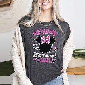 May include: Dark grey t-shirt with the text "MOMMY OF THE BIRTHDAY GIRL" in pink and white. The design features a black Minnie Mouse head with a pink bow and stars. A fun and festive shirt for a birthday celebration.