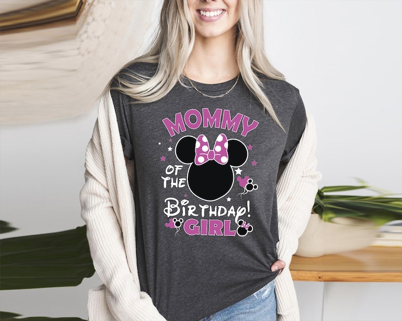 May include: Dark grey t-shirt with the text "MOMMY OF THE BIRTHDAY GIRL" in pink and white. The design features a black Minnie Mouse head with a pink bow and stars. A fun and festive shirt for a birthday celebration.
