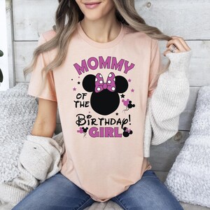 May include: A peach-coloured t-shirt featuring the text "Mommy of the Birthday Girl" in purple and black, with a Minnie Mouse design. The design includes a polka-dot bow. The shirt is a casual, short-sleeved top.
