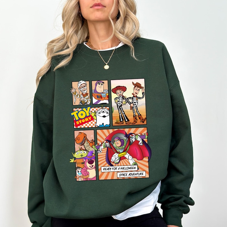 May include: Dark green sweatshirt featuring a colorful comic strip design with characters from the Toy Story movie. The comic strip includes images of Woody, Buzz Lightyear, and other characters. Text on the sweatshirt reads "TOY STORY" and "READY FOR A HALLOWEEN SPACE ADVENTURE."