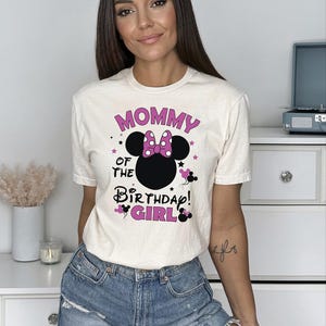 May include: Cream-coloured t-shirt with the text "MOMMY OF THE BIRTHDAY GIRL!" in pink and black. The design includes a Minnie Mouse silhouette with a pink polka-dot bow. The shirt is paired with denim shorts.